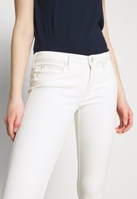 Pantalones de mezclilla blancos de corte ajustado, con bolsillos frontales, cierre de botón y trabillas para cinturón. Tejido texturizado con un ligero estiramiento.