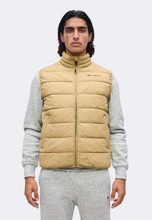 Homme aux longs cheveux foncés portant un gilet matelassé beige par-dessus un sweat-shirt gris clair et un pantalon de jogging, face à l’avant avec une expression neutre.