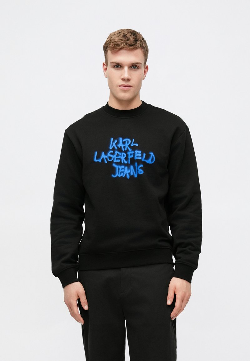 Svart genser med lange ermer og ribbede mansjetter. Har en fremtredende blå spraymalt logo som leser "Karl Lagerfeld Jeans" på forsiden.