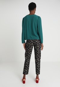 Žena s krátkými kudrnatými vlasy má na sobě tyrkysový top a kalhoty s zvířecím potiskem, stojí zády k fotografovi v minimalistickém prostoru.