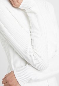 Biała kurtka o teksturowym wzorze z żebrowanym wzorem, z dopasowanymi rękawami i przytulnym kołnierzem. Materiał jest miękki i lekko elastyczny.