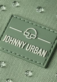 Tessuto verde con una toppa rettangolare cucita etichettata "JOHNNY URBAN" e un volto minimalista con occhiali sopra il testo.