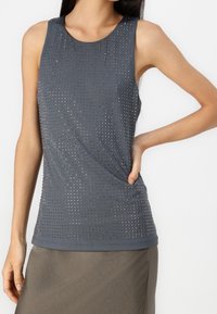 Top sin mangas de color gris oscuro con pequeñas tachuelas metálicas, llevado por una persona con la mano en la cadera, combinado con una falda marrón.