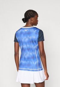 Blå og svart treningsbluse med gradientdesign, korte ermer og kontrastsøm, satt sammen med et hvitt perforert skjørt.