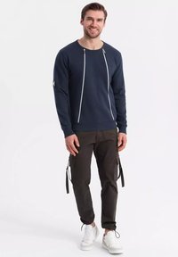 Marinblå sweatshirt med vita dragkedjedetaljer, rund halsringning; tillsammans med svarta cargobyxor med sidofickor och vita sneakers.