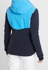 Modrá a námořnicky modrá lyžařská bunda s pevnou kapucí, dlouhými rukávy a černými detaily. Hladká textura látky s kontrastními barevnými částmi.