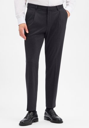Mann trägt dunkelgraue maßgeschneiderte Hose, schwarze Anzugsschuhe, schwarze Socken und ein weißes Hemd mit einer Hand in der Tasche.