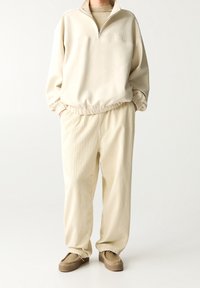 Beige kwart-zip sweater met elastische manchetten, gecombineerd met textuurbeige corduroy broeken. Model draagt bruine instap schoenen.