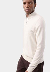 Cremefarvet strikpullover med halvlynlås krave, ribbede manchetter og taljebånd, parret med mørkebrune bukser; glat tekstur og afslappet design.