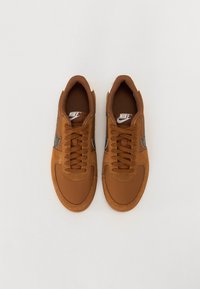 Sapatilhas em camurça e malha castanhas com bico arredondado, atacadores planos e um logo em forma de swoosh cinza nas laterais. Detalhes brancos no calcanhar.