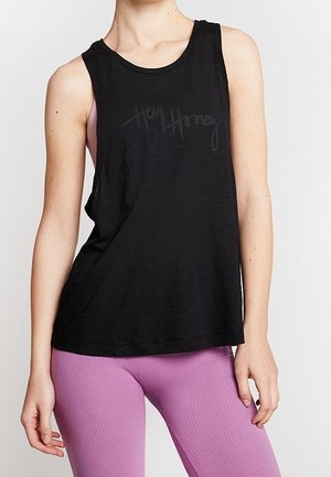 Frau in einem schwarzen ärmellosen Oberteil mit der Aufschrift "Hey Honey" und rosa gerippten Leggings, dargestellt vom Hals bis zur Hüfte vor einem einfarbigen Hintergrund.