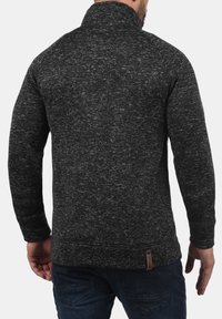 Miesten tummanharmaa poolopaita, jossa kohotettu kaulus, teksturoidulla neulekuviolla ja nahkainen korostelappu helmassa.