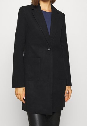 Frau trägt einen langen schwarzen Mantel mit einem Knopf, Lapellen und aufgesetzten Taschen, kombiniert mit schwarzen Lederhosen und einem blauen Oberteil.