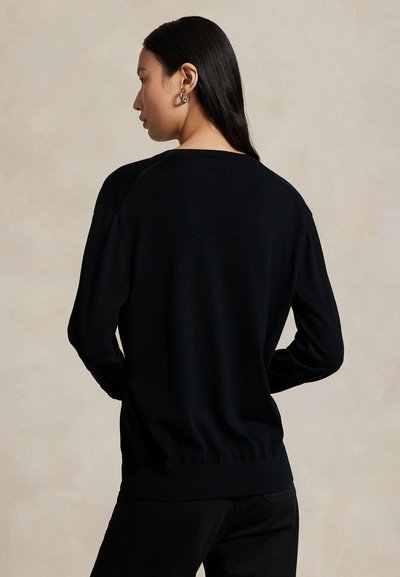 Woman with long black hair wearing a black long-sleeve sweater and black pants, facing sideways against a plain beige background.