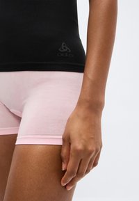 Κοντινό πλάνο από τον κορμό και το πάνω μέρος των ποδιών ενός ατόμου που φορά μαύρο τοπ και ανοιχτό ροζ στενά σορτς, με το δεξί χέρι χαλαρό δίπλα στο πλευρό.