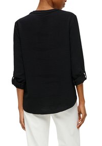 Haut noir avec un tissu texturé, un col rond et des manches 3/4 à revers. Le dos présente un ourlet arrondi et est légèrement plus long.