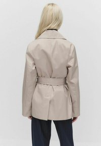 Beżowy płaszcz trench z dopasowanym pasem, długimi rękawami i szczegółem wentylacyjnym z tyłu. Gładki, teksturowany materiał. Zwróć uwagę na czysty krój i wyrafinowany design.