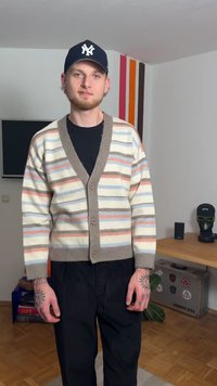 Gestreifter Cardigan in Creme, mit braunen, pinken und blauen horizontalen Streifen. Strickstruktur, V-Ausschnitt und vier Knöpfe. Kombiniert mit schwarzen Hosen.