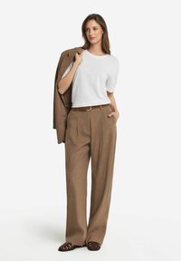 Hvid t-shirt parret med brune plisserede bredbenede bukser og en brun blazer draperet over den ene skulder. Brun bælte og mørke sandaler fuldender looket.