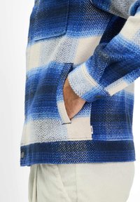 Giacca a quadri blu con texture a spina di pesce in diverse tonalità di blu e bianco, dotata di tasca e polsini con bottoni.