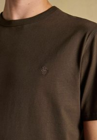 Brūns kreklu ar apaļu kakla izgriezumu un īsām piedurknēm, ar mazu izšūtu logotipu kreisajā krūtī, ko valkā cilvēks.