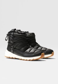 Bottes d'hiver noires isolantes montantes jusqu'à la cheville, avec semelles épaisses blanches, lacets noirs à motifs, et semelles extérieures en caoutchouc couleur tan sur un fond uni.