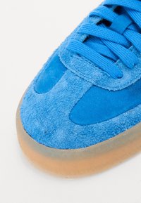 Modré semišové tenisky s texturovaným svrškem, hladkými akcenty a kulatou špičkou. Mají modré tkaničky a průhlednou gumu na podrážce.