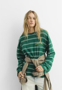 Ung kvinna med långt blont hår som bär en grönrandig långärmad skjorta med text, en beige jacka knuten runt midjan och ljusa denimjeans.