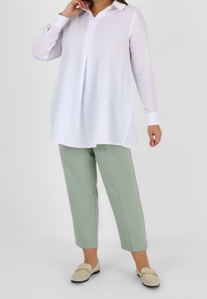 Femme portant une blouse blanche à manches longues ample, un pantalon court vert sauge et des mocassins beiges avec un détail de chaîne dorée, debout devant un fond blanc.