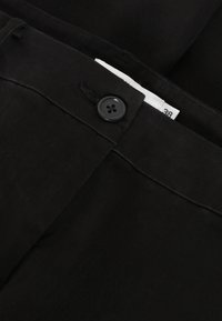 Schwarze Hose aus glattem Stoff mit Knopfverschluss und einem sichtbaren Etikett am Bund. Der Knopf hat vier Löcher zur Befestigung.