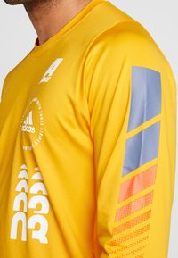 Gelbes Sport-T-Shirt mit langen Ärmeln, mit rundem Ausschnitt, weißem Adidas-Logo und farblich abgestimmten grauen und roten Akzenten an den Ärmeln.