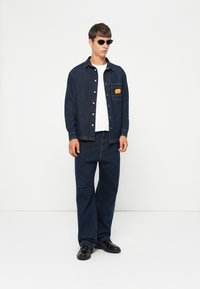 Dark blue denim jacket with a chest pocket, paired with matching loose-fitting jeans. Worn over a white t-shirt; black sunglasses complete the look.