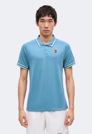Jonge man die een lichtblauw poloshirt met korte mouwen draagt, met een witte bies en een klein meerkleurig logo op de borst, gecombineerd met witte shorts.