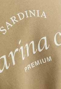 Beige fabric with white printed text: "SARDINIA" curved above "marina c" and "PREMIUM" below. Smooth texture, slightly elastic appearance.