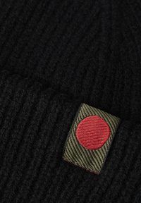 Mustra mustra kootud beanie, millel on tekstuuriga roheline silt ja silmapaistev punane ring. Materjal näib olevat pehme ja soe.