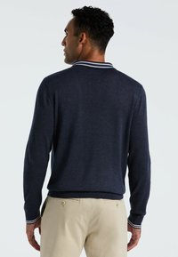 Dunkelblaues Strick-Polohemd mit einem weißen Akzent am Kragen. Lange Ärmel und gerippte Bündchen. Modell trägt beigefarbene Hose.