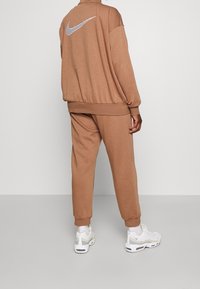 Brun idrottsoverall med lös passform. Har en stor vit Nike-logotyp på ryggen och kombineras med vita sneakers.