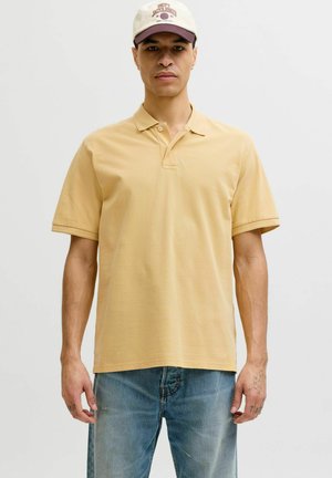 Homme portant un polo beige, un jean bleu et une casquette de baseball blanche et bordeaux, debout devant un fond uni.