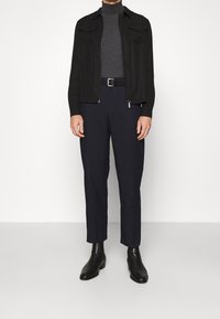 Veste zippée noire avec deux poches poitrine, col roulé gris, pantalon bleu foncé et bottines en cuir noir. Tissu lisse, coupe ajustée.