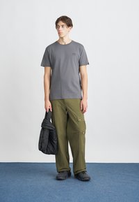 T-shirt de algodão cinza com mangas curtas e um pequeno logotipo, combinada com calças cargo verde-oliva feitas de material leve. Casaco preto isolante pendurado.