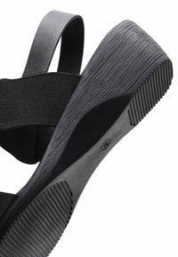 Sandale compensée noire montrant une semelle en caoutchouc texturée avec des rainures, une bride élastique à la cheville et une bride arrière lisse au talon.