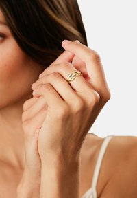 Goldring mit einem geflochtenen Design aus glatten und strukturierten Gliedern, getragen an einer Hand mit hellerer Hautfarbe vor einem neutralen Hintergrund.