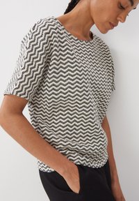 Pull en maille crème et noir avec motif en zigzag, manches courtes, encolure ronde et coupe décontractée. Détail de tissu texturé visible.