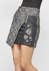Jupe en denim avec une taille texturée à motif en chevrons et des détails floraux brodés à l'avant. Longueur courte, tissu à teinte foncée.