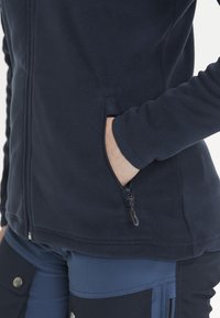 Veste en polaire navy avec poche zippée sur le côté, texture douce, design ajusté. Les bras sont ajustés, avec un ourlet inférieur sans coutures pour un confort de mouvement.