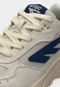 Αθλητικό παπούτσι ανοιχτό γκρι με υλικά από δίχτυ και δέρμα σουέτ. Διαθέτει μπλε λογότυπο, επίπεδα κορδόνια και υφή στην εξωτερική σόλα.