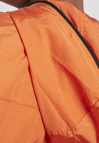 Orange letvægtsjakke med en glat tekstur, synlige sømdetaljer og en sort lynlås langs hætten og skulderen.