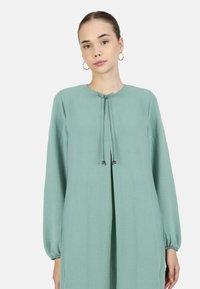 Long-sleeve, light green dress with a rounded neckline, tie closure, and textured fabric. Features relaxed fit and gathered sleeve cuffs.