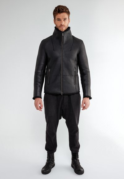 Uomo che indossa una giacca nera aderente con zip e colletto alto, pantaloni scuri larghi e scarpe sportive nere alte, in piedi contro uno sfondo semplice.