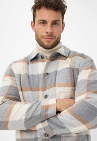 Veste-chemise à carreaux dans des tons gris, blanc et beige. Fabriquée en tissu doux, elle comporte une fermeture à boutons sur le devant et une poche poitrine. Finition texturée.